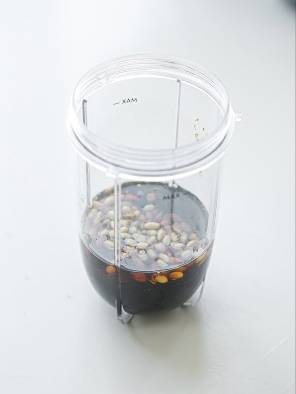 A clear blender cup containing soy sauce and whole peppercorns sits on a light-colored surface, viewed from above.